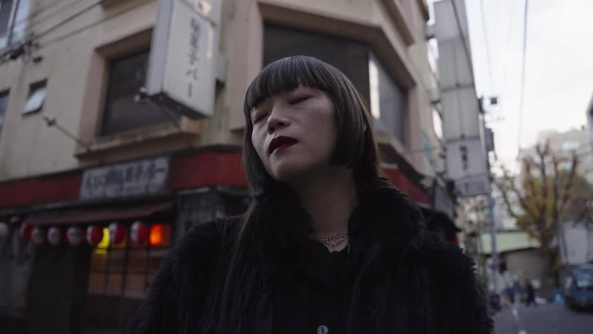 A stylish young Japanese woman in a black fur coat gazes contemplatively on a quiet city street in slow motion