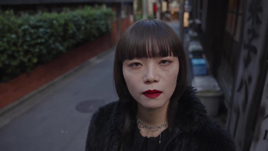 A stylish young Japanese woman with red lipstick stares intensely at the camera while standing in a narrow city alleyway