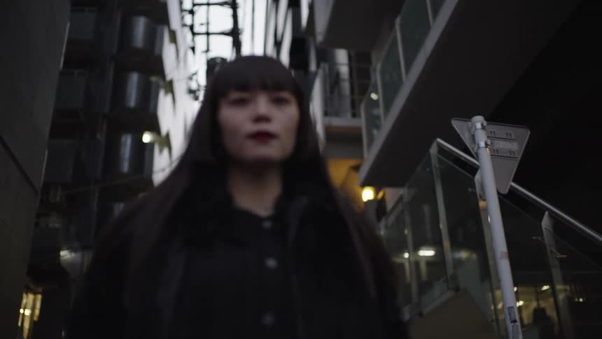 A stylish Japanese woman in all black walks toward the camera in slow motion down a dimly lit city alleyway