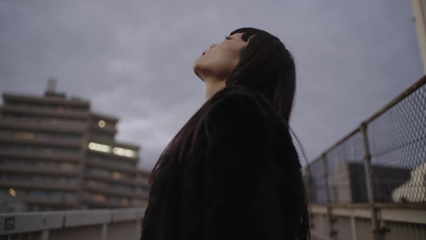A thoughtful young Asian woman in a black coat stands on an urban footbridge gazing up at the overcast sky