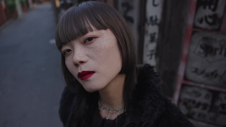 Close-up slow-motion shot of a stylish Asian woman with red lipstick and bangs staring intensely at the camera in a narrow urban alley