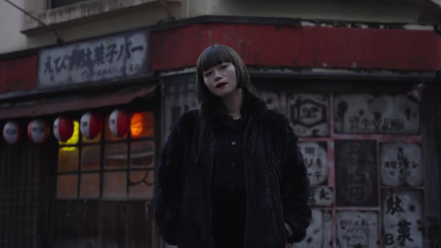 A chic Japanese woman in a black fur coat poses for a portrait on a city street in front of a traditional storefront with glowing lanterns