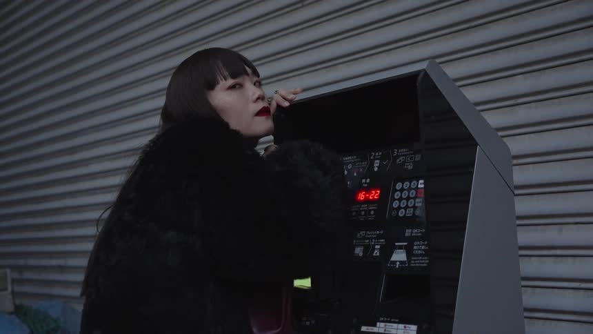 A stylish and sad Japanese woman in a black fur coat stands pensively at a public payphone in a quiet city street