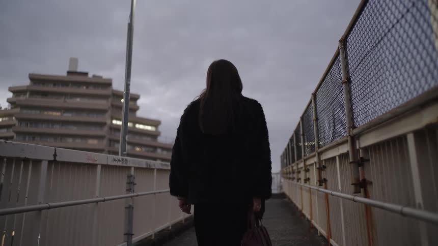 A stylish woman in all black walks down a fenced-in city walkway at twilight, briefly looking back over her shoulder in slow motion