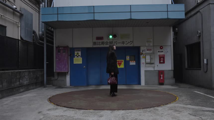 A fashionable woman in an all-black outfit stands on a slowly rotating vehicle turntable in an urban setting