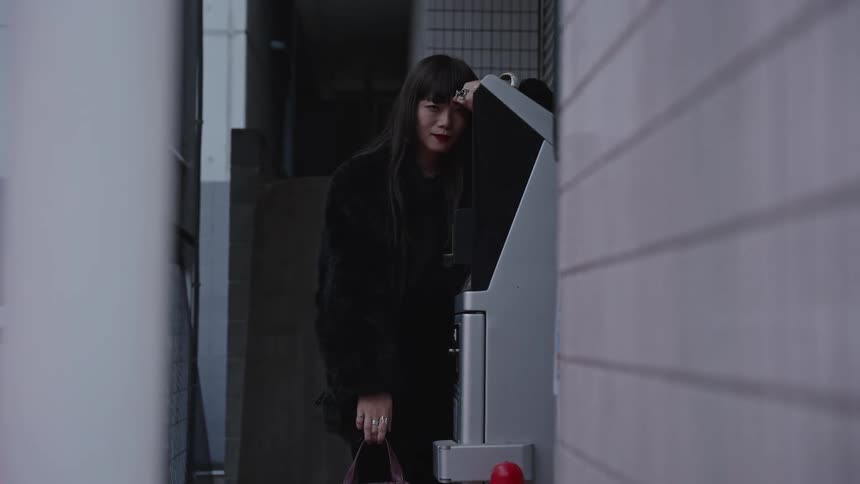 A stylish woman in a black fur coat poses against a parking meter in an urban alleyway, looking directly into the camera