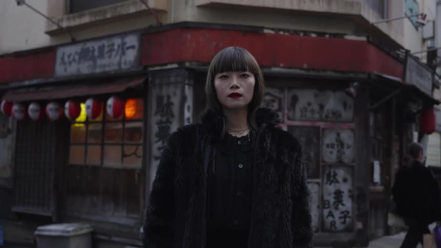 A stylish woman in a black fur coat stares intently at the camera on a moody, traditional Japanese street
