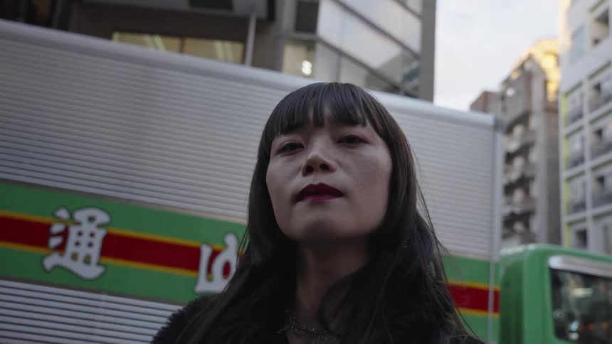 A fashionable Asian woman stares intently into the camera in a dramatic low-angle slow-motion shot on a city street