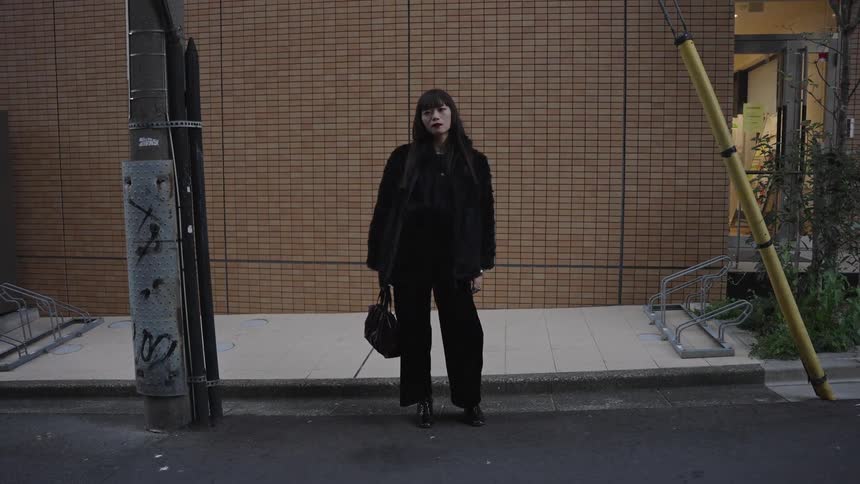 A stylish woman in all black poses on a city street, looking directly at the camera as it slowly zooms out
