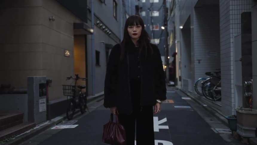 A stylish woman in a black outfit stands still and looks directly at the camera in a narrow city alley at dusk