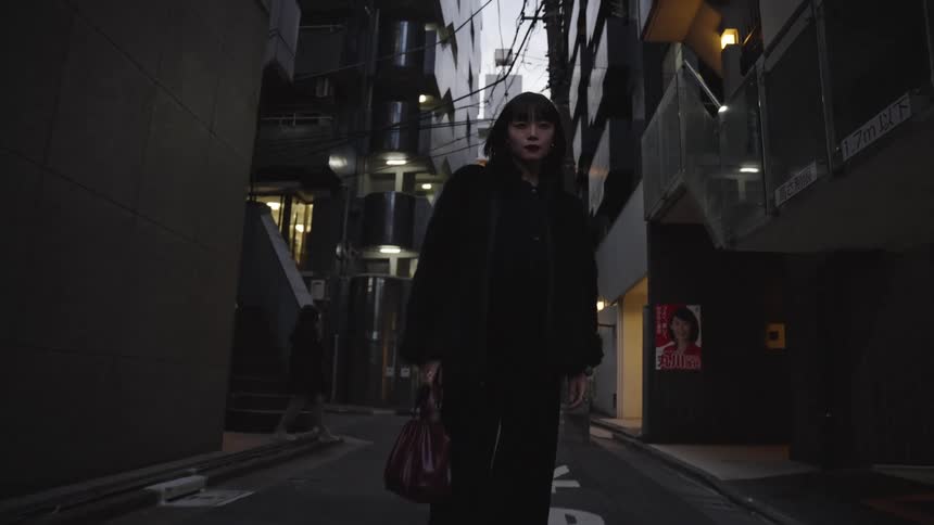 A stylish young woman in a black coat stands in a dark urban alleyway in Tokyo, looking at the camera as it zooms in