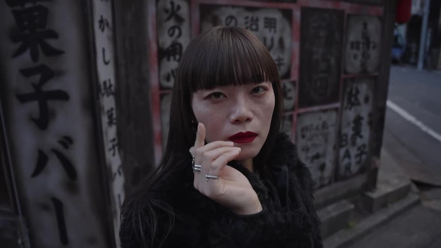 An enigmatic Asian woman with bold red lipstick and blunt bangs gazes intensely at the camera on a moody city street in slow motion