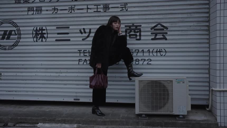 A fashionable woman in a black fur coat strikes a pose on a city sidewalk in Japan, resting her foot on an air conditioning unit