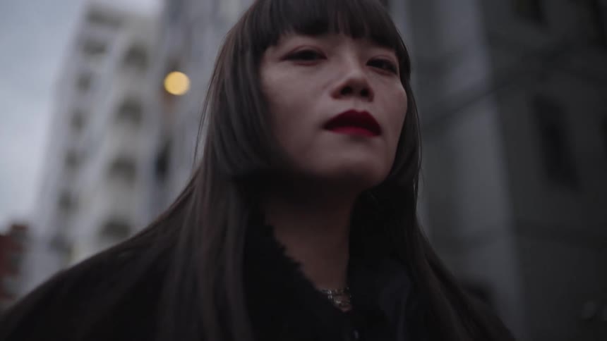 An enigmatic young woman with an intense gaze walks through a moody city street in dramatic slow motion
