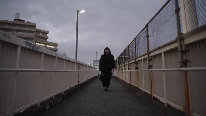A stylish woman in all black walks toward the camera in slow motion on a gritty urban footbridge at dusk