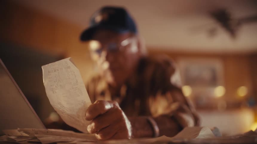 A close-up shot focuses on an elderly man's hand holding a receipt as he thoughtfully reviews his finances at a table
