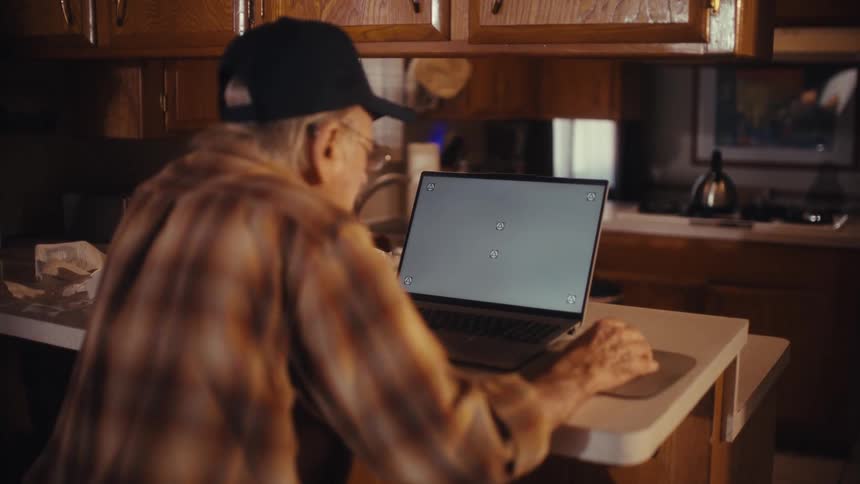 An elderly man wearing a plaid shirt and baseball cap uses a laptop with a green screen at his kitchen counter