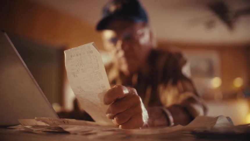 A senior citizen wearing a baseball cap and glasses checks his expenses by reading a long receipt at his desk
