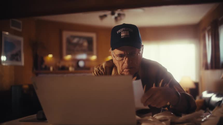 Slow zoom in on an elderly retired firefighter carefully reviewing his bills and using a laptop to manage his finances at home