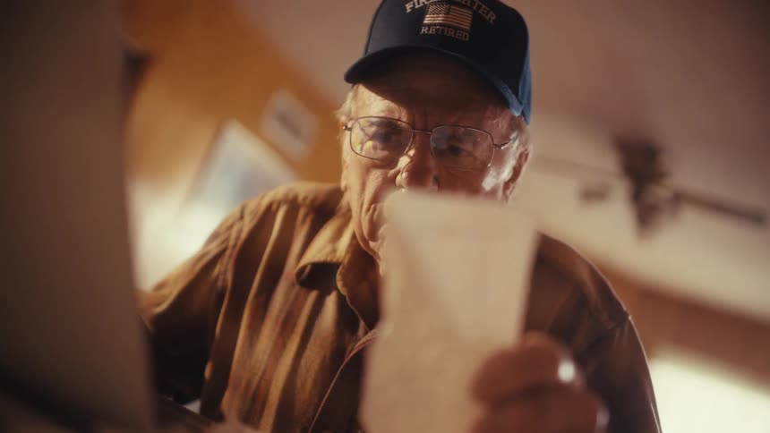 An elderly retired firefighter in a cap and glasses intently reviews bills while using a laptop at his desk