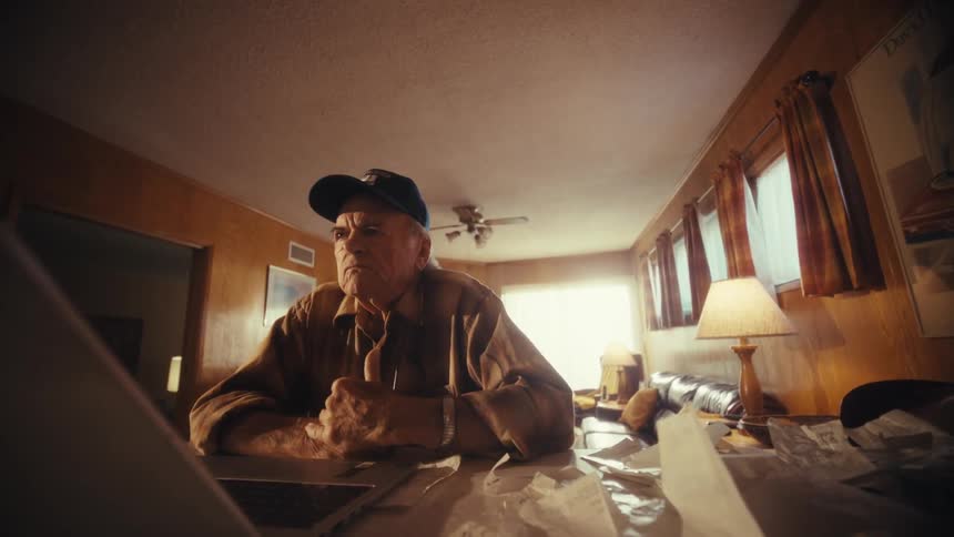 A pensive elderly man wearing a baseball cap sits at a cluttered table looking at his laptop, possibly stressed about finances