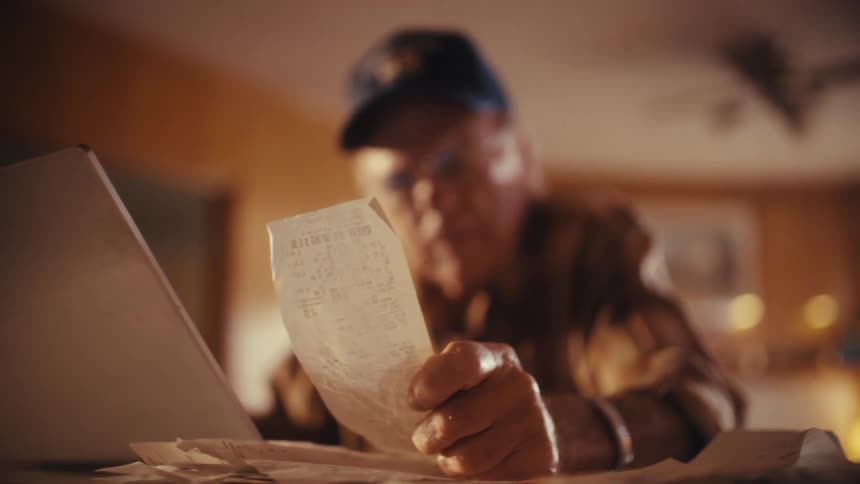 An elderly man sits at his kitchen table and reviews his monthly expenses by looking over his receipts