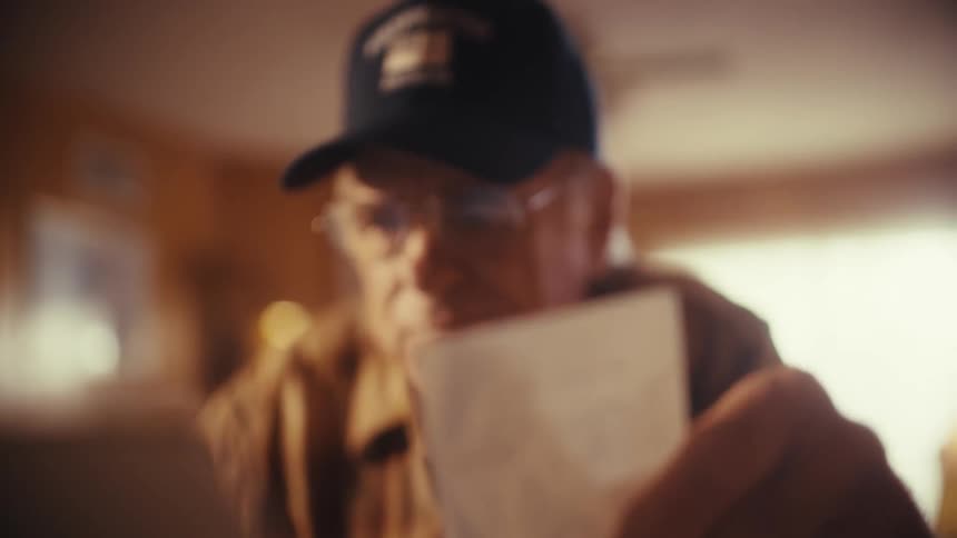 A retired firefighter wearing glasses and a hat carefully reviews a crumpled receipt at his sunlit table