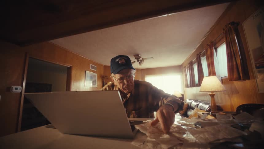 A senior man with a hat and glasses carefully reviews receipts while managing his finances on a laptop in his home