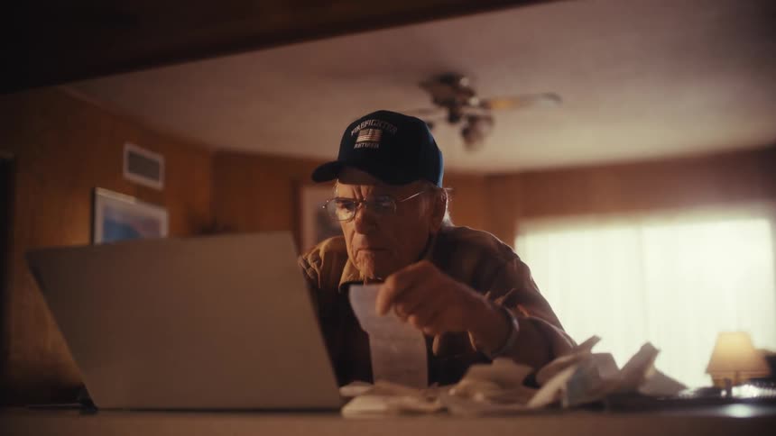 An elderly retired firefighter carefully reviews his bills and receipts on his laptop while managing his finances at home