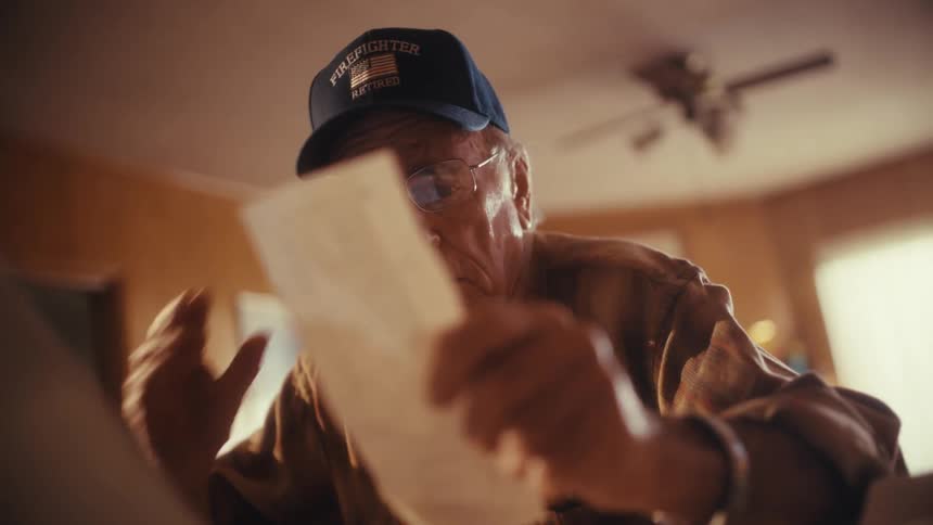 A worried elderly man wearing a retired firefighter hat becomes stressed and shocked while reviewing his bills and finances at home