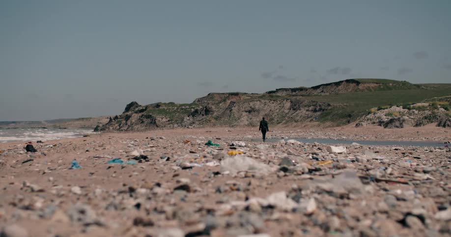A solitary person walks away from the camera across a sandy beach completely covered in trash and plastic pollution