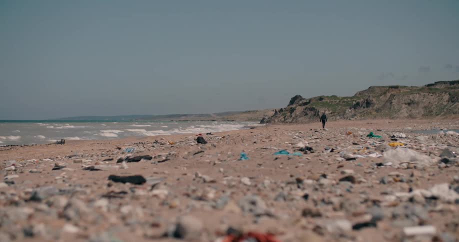 A low-angle, long shot shows a lone figure walking along a sandy beach completely covered in plastic pollution and garbage