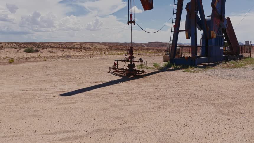 A slow pull-out reveals an oil pumpjack operating in the vast, arid desert under a cloudy sky