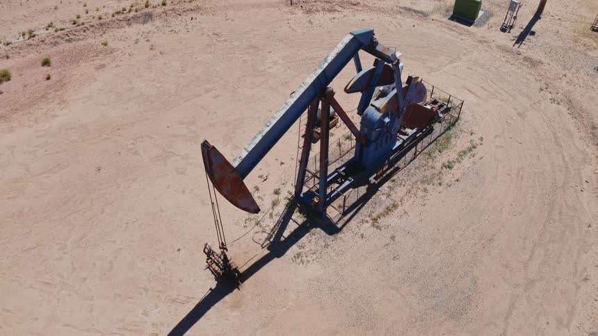 An aerial view of a rusty oil pump jack rhythmically extracting crude oil from the arid desert ground on a sunny day