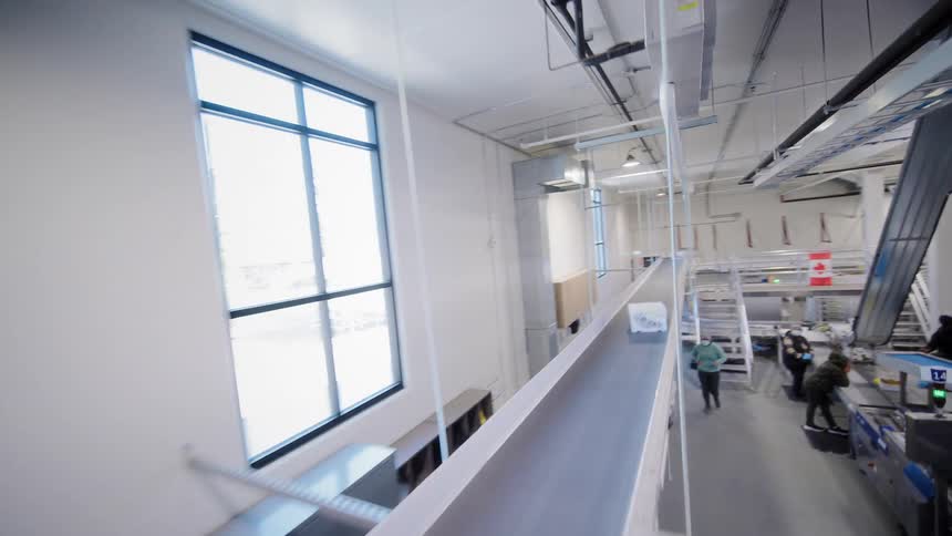 A high-angle tracking shot reveals the vast interior of a modern food processing facility with intricate conveyor belt systems and workers