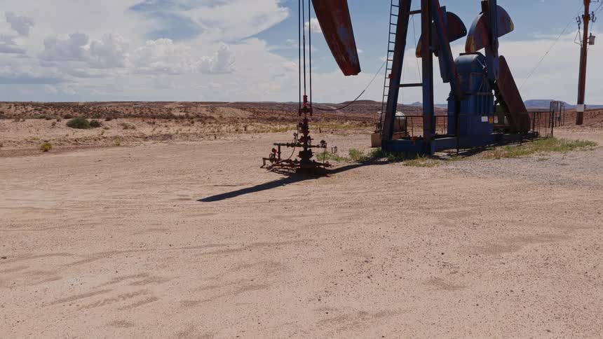 A stationary oil pump jack stands idle in a vast, arid desert landscape under a partly cloudy sky