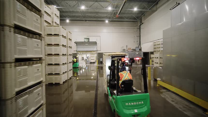 Smooth gimbal shot follows a forklift driver transporting large white crates through a brightly lit, wet industrial food processing warehouse