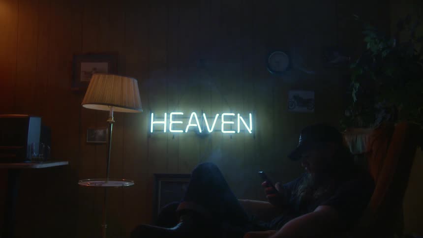 Man sits on a couch in dim room with neon sign