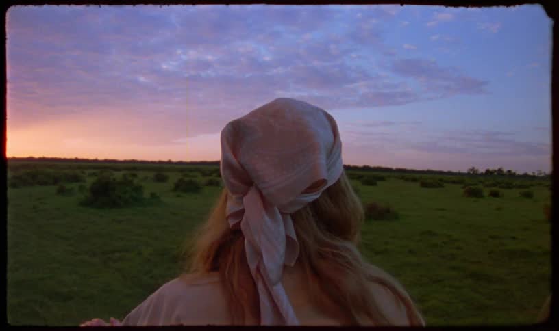 A woman wearing a headscarf looks out over a vast green field during a colorful sunrise, captured on vintage film