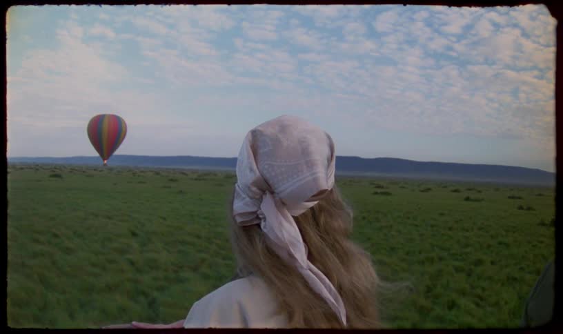 A blonde woman with a headscarf enjoys a serene hot air balloon ride, watching another colorful balloon drift over a vast green savanna