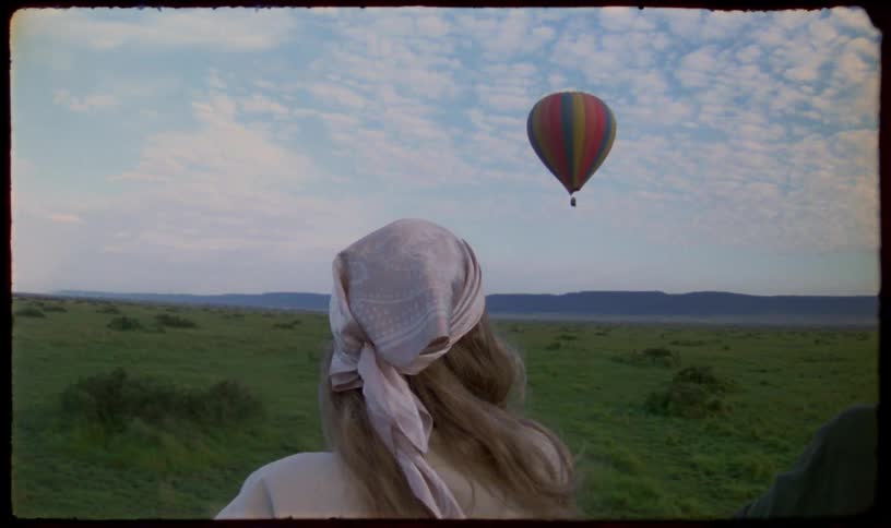 Over-the-shoulder shot of a woman watching a colorful hot air balloon float over a vast green savanna