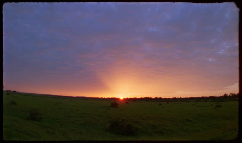 A vintage, wide-angle shot shows a serene sunset over a vast, green field under a purple and orange sky