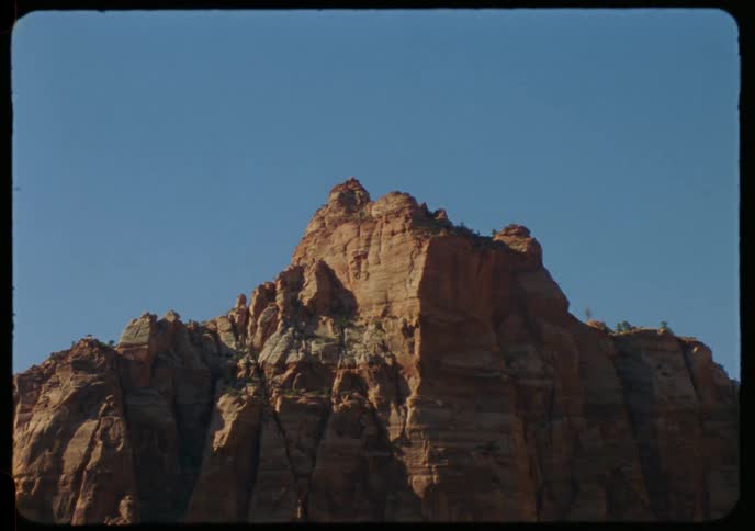 A vintage, static shot captures the rugged peak of a sunlit red rock mountain against a vast, clear blue sky