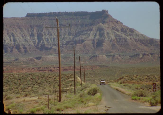 A vintage white pickup truck drives towards the camera on a remote desert road with large mesas in the background