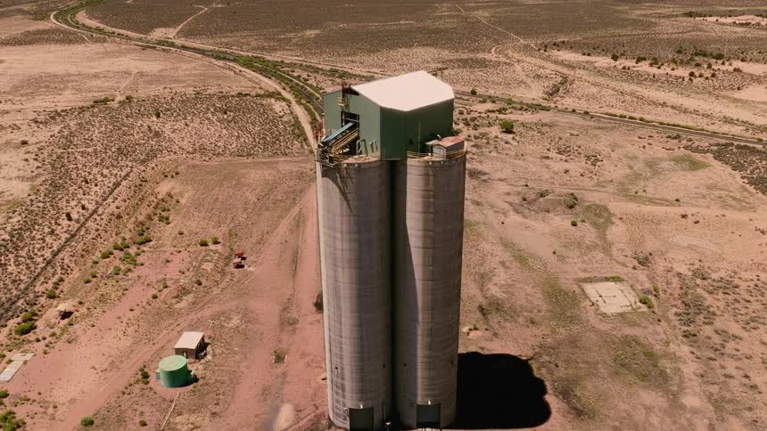An aerial view slowly pulls back from a tall concrete grain silo in the middle of a vast, arid desert landscape