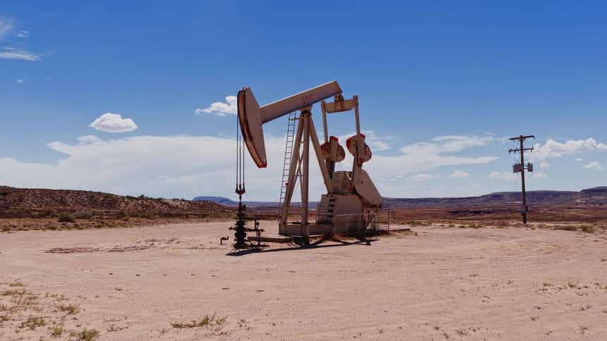 A large oil pumpjack rhythmically works to extract crude oil from a well in the middle of an arid desert on a sunny day