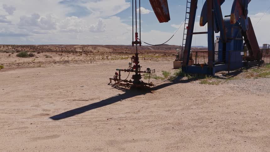 A dolly shot moves towards the wellhead of an oil pumpjack operating in the vast, arid desert under a partly cloudy sky