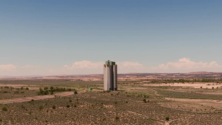 Aerial view slowly pulls back from a tall grain silo standing alone in a vast, arid desert landscape under a clear blue sky