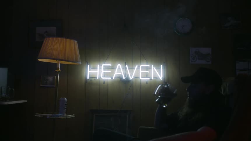 Man sits drinking on a couch in a dim room light with a neon sign
