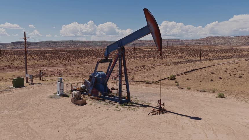 An oil pumpjack operates continuously in a vast, arid desert under a bright blue sky